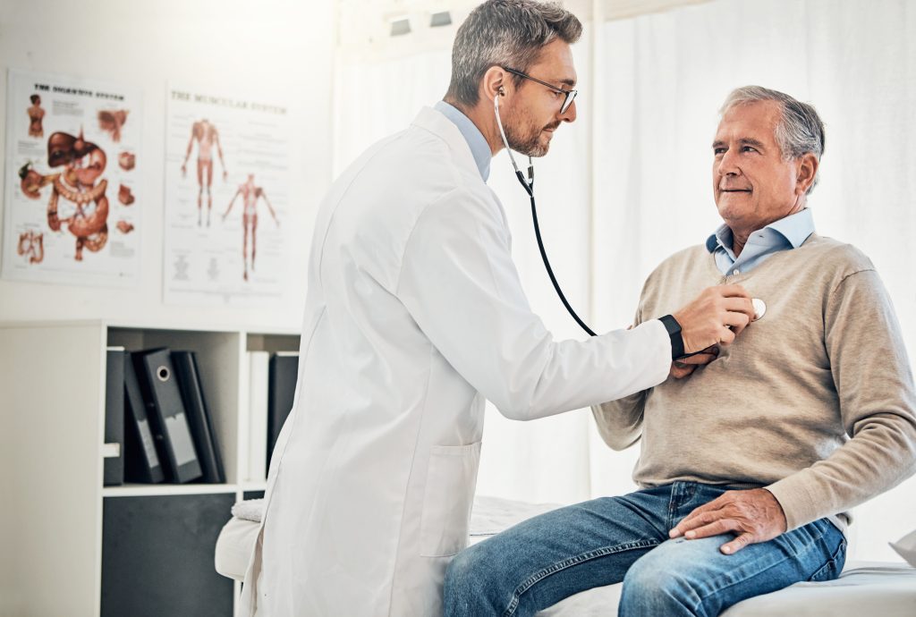 Senior white male patient sitting on exam table in doctor examination room. The tall white male doctor holds a stethoscope to the patient's chest.
